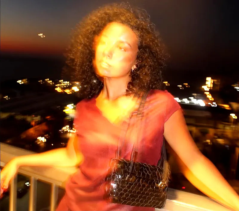 Blurred night rooftop portrait with a black woven shoulder bag, city lights below